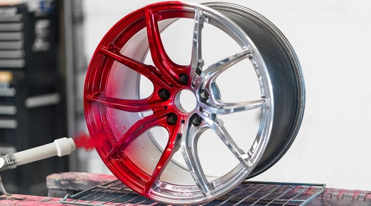 Car wheel rim being half-painted red with a spray gun, half remains silver.