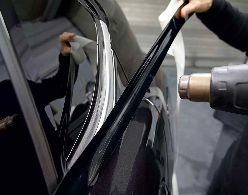 Person applying black vinyl wrap to the window frame of a car using a heat gun.