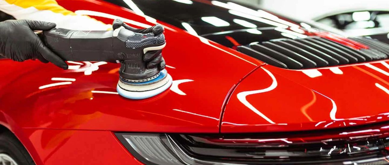 Close-up of a red car being polished with an electric buffer by a gloved hand.