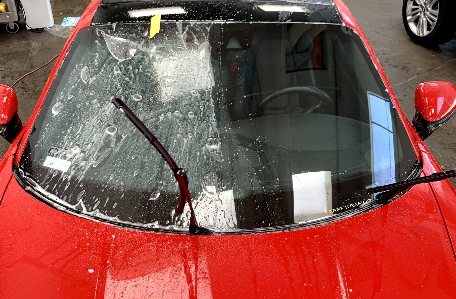 Red car with wet windshield, wipers lifted, and interior partially visible in a garage setting.